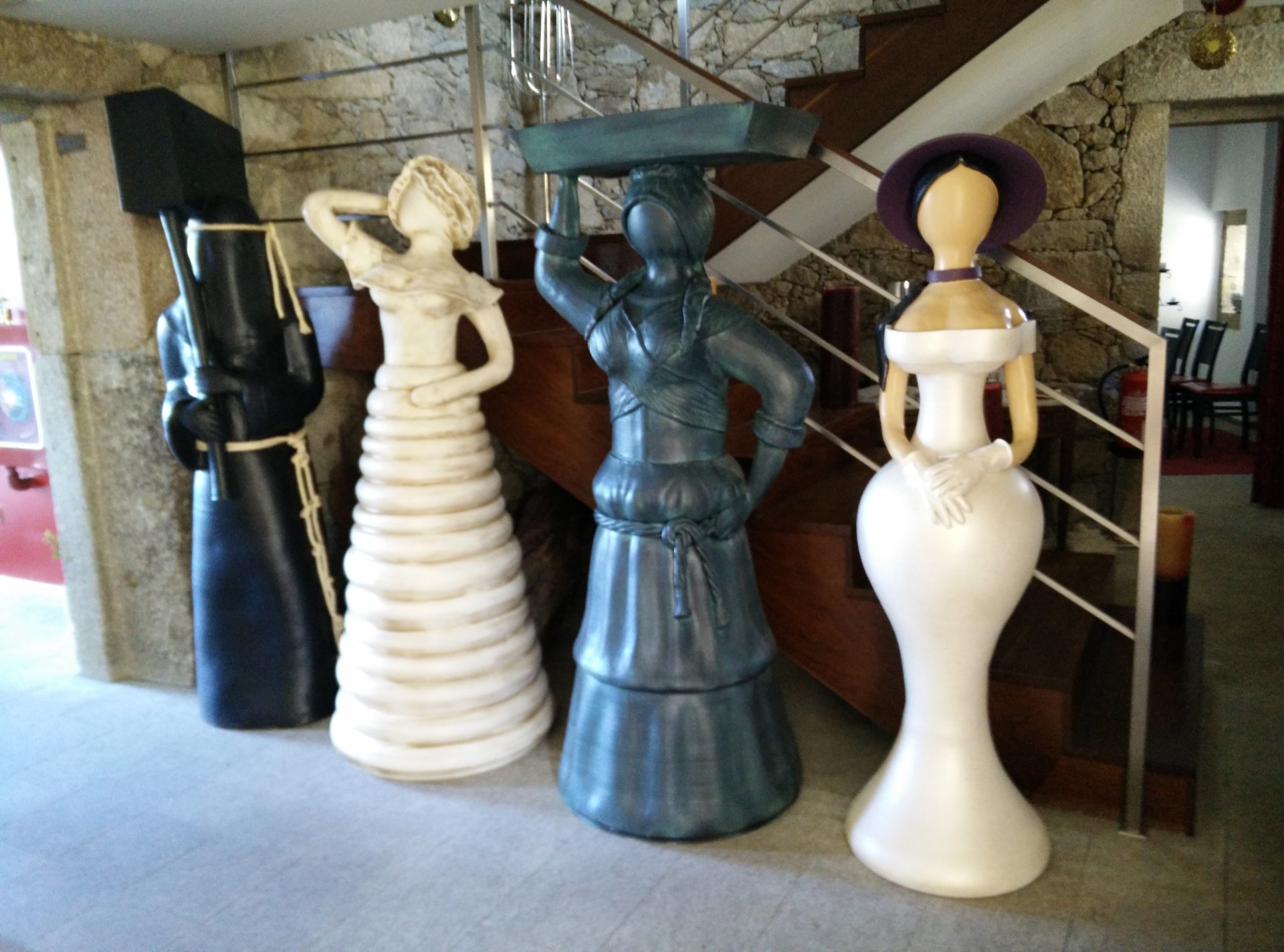 Quatro esculturas femininas grandes em materiais variados junto a escadas interiores.