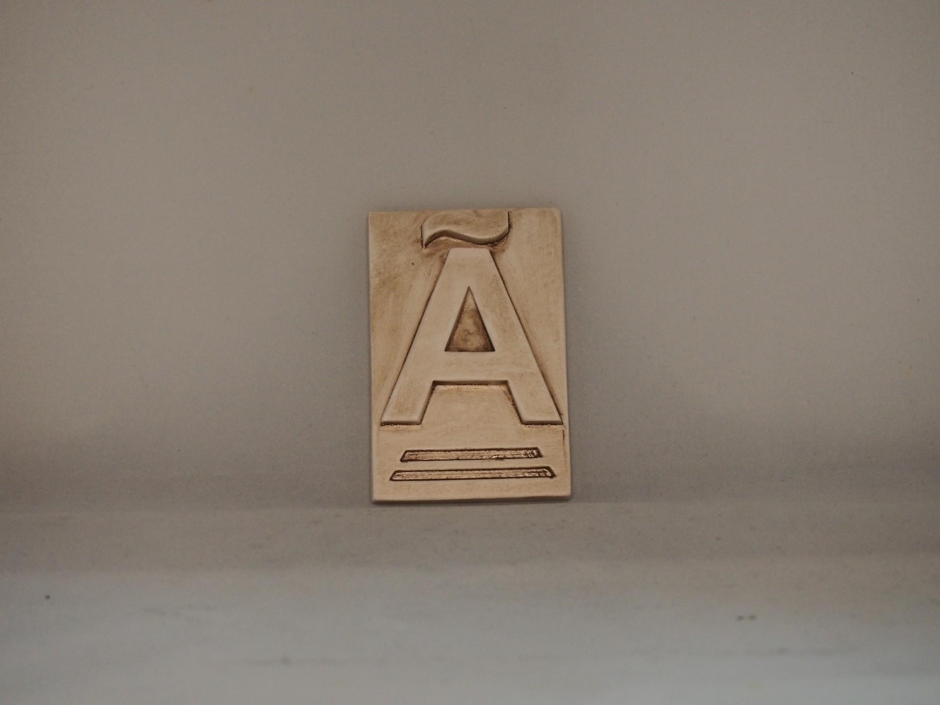 Placa de madeira com letra Á em relevo sobre fundo cinzento