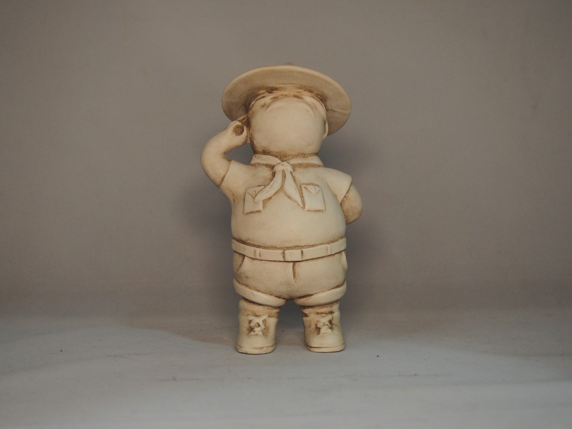 Figura decorativa de criança vestida com chapéu, lenço, camisa, calções e botas em tom bege.