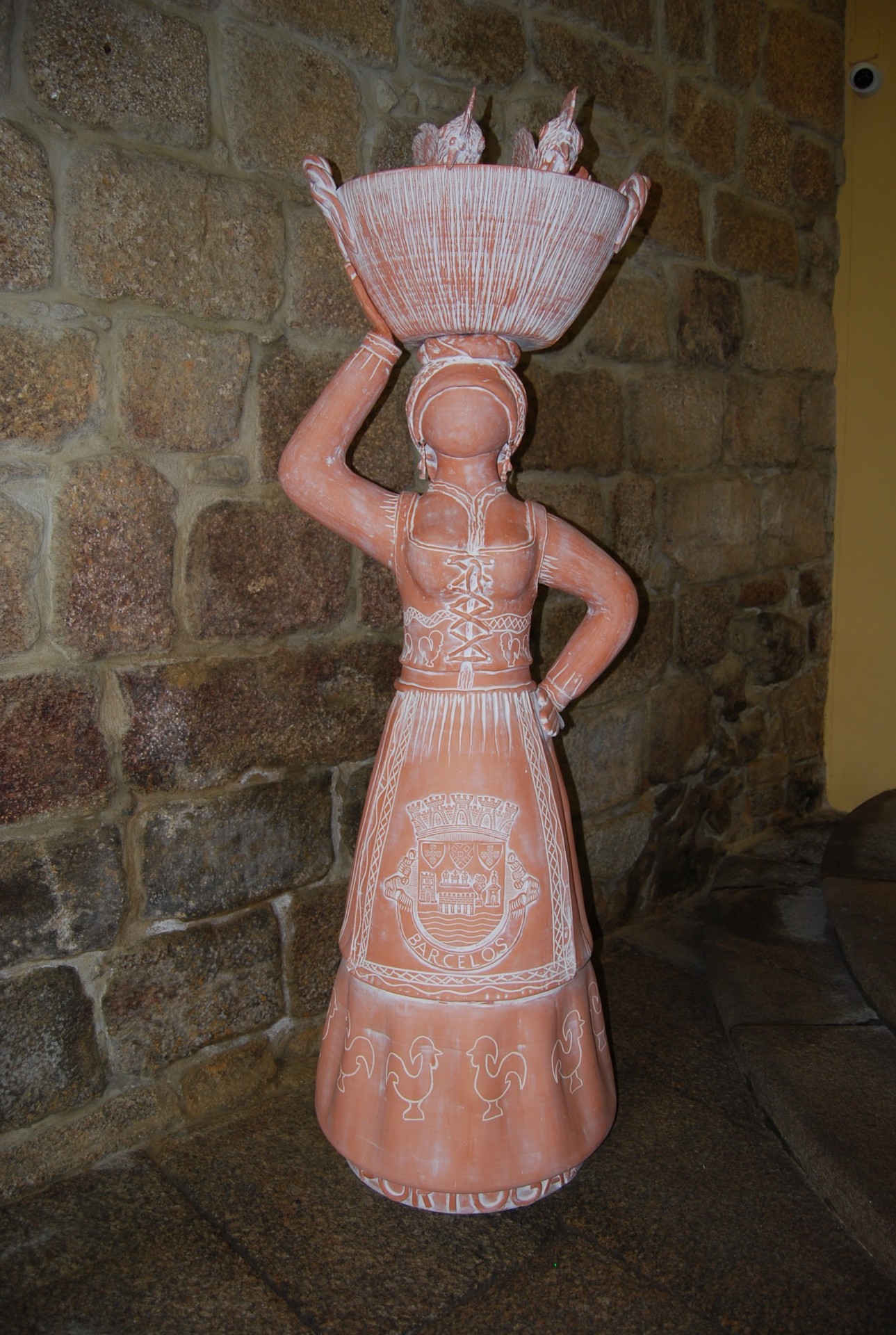 Estátua de mulher em terracota segurando tigela com galos, com brasão de Barcelos na saia.