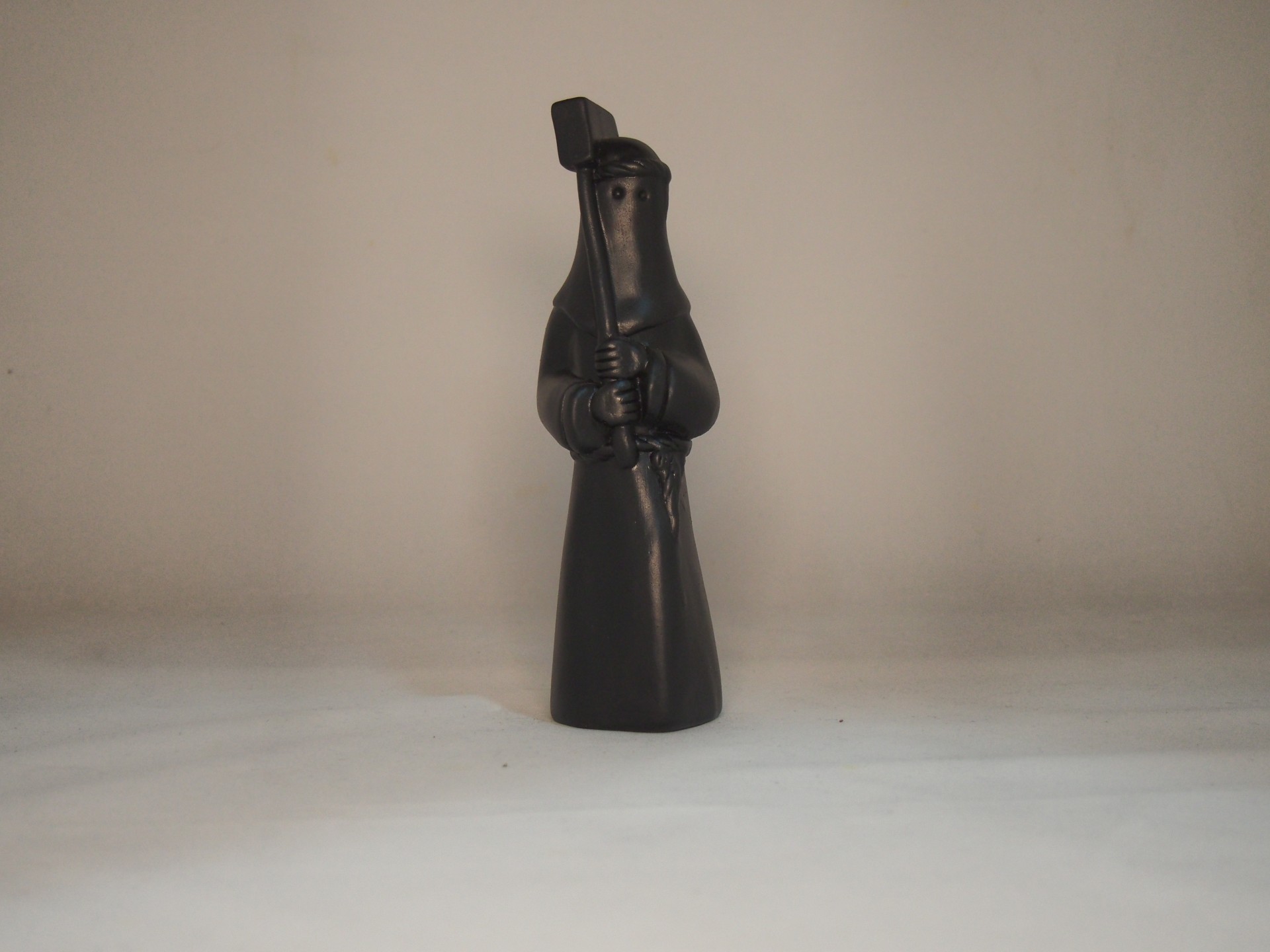 Figura decorativa preta com túnica longa e capuz segura objeto alongado em fundo neutro
