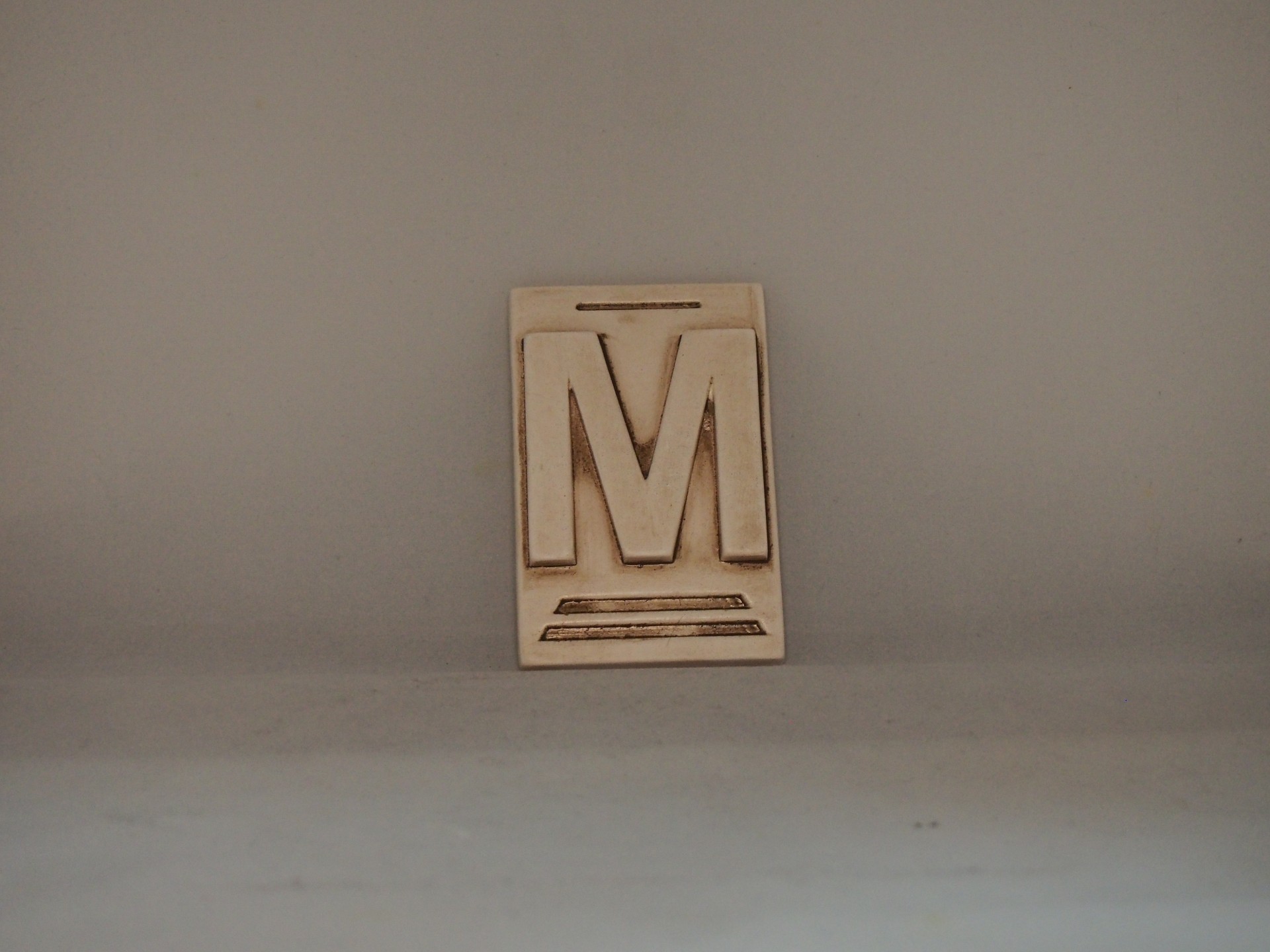 Peça decorativa em madeira com letra M em relevo.
