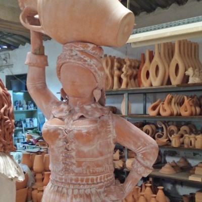 Escultura em terracota de mulher com jarro na cabeça e várias peças de cerâmica ao fundo