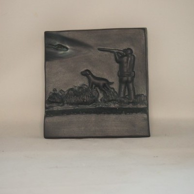 Placa decorativa em metal escuro com relevo de caçador e cão.