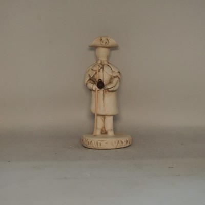 Figura decorativa bege com chapéu e bengala sobre base redonda em fundo cinzento.