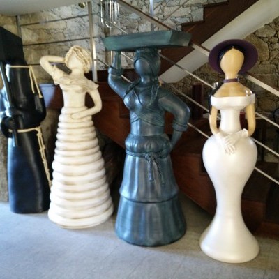 Quatro esculturas femininas grandes em materiais variados junto a escadas interiores.