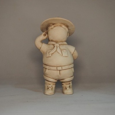 Figura decorativa de criança vestida com chapéu, lenço, camisa, calções e botas em tom bege.