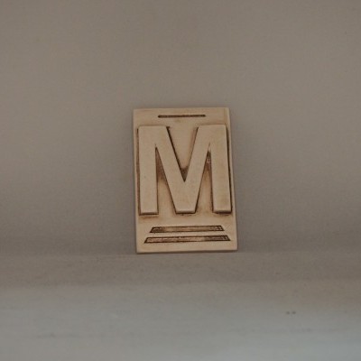 Peça decorativa em madeira com letra M em relevo.