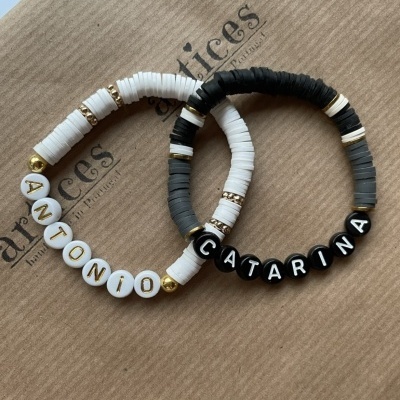Duas pulseiras customizadas com discos coloridos e contas com nomes ANTONIO e CATARINA sobre papel castanho com texto ARTices