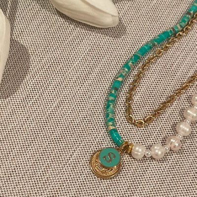 Colar com pendente 'S' em verde e dourado, pérolas brancas e corrente dourada sobre tecido bege