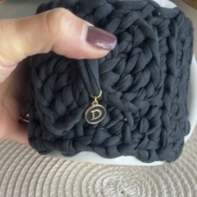 Bolsa de malha preta trançada com pendente letra D, segurada por mão com unhas castanho-escuro