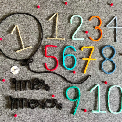 Números coloridos em lã sobre fundo cinzento com pontos vermelhos e palavras em fio preto.