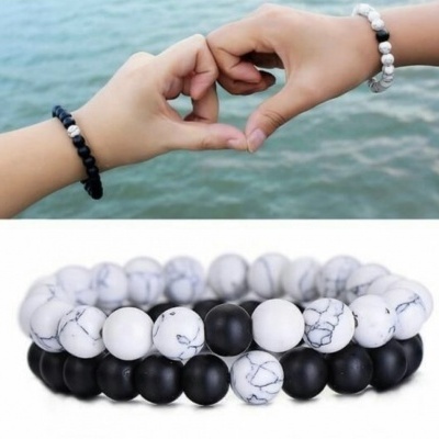 Duas pulseiras de contas, uma branca com padrão de mármore e outra preta fosca, em pulso de duas pessoas formando um coração com as mãos.