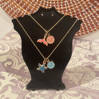 Dois colares dourados com pingentes e medalhões com a letra M expostos num suporte preto contra fundo com padrão geométrico