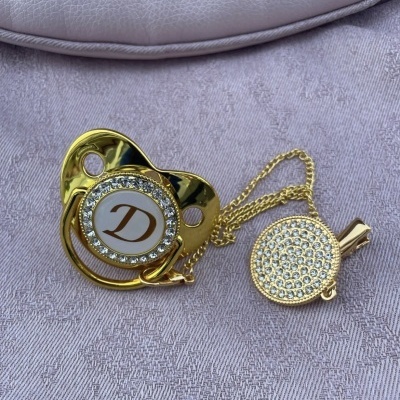 Chupeta decorativa dourada com letra D e corrente com prendedor dourado cristais sobre tecido lilás