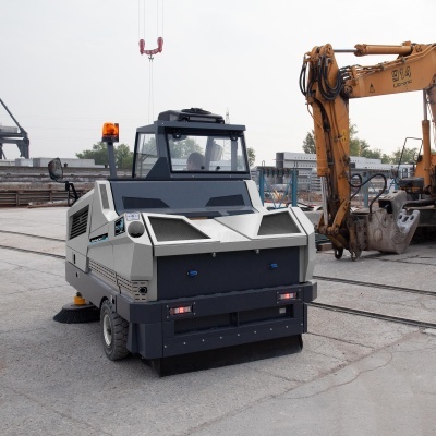 Varredora industrial cinza e preto numa área industrial com escavadora amarela ao fundo