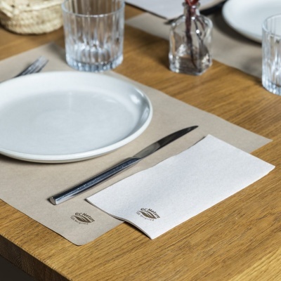 Guardanapo branco e individual castanho com logo ECOMIC, prato branco, faca e copo em mesa de madeira