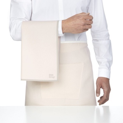 Pano de cozinha creme pendurado no braço de pessoa com camisa branca e avental creme com bolso