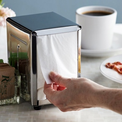 Guardanapos de papel brancos numa caixa metálica e mão a puxar um guardanapo numa mesa com café e torta ao fundo