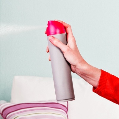 Mão com manga vermelha a segurar spray cinzento com tampa rosa a ser usado em ambiente interior