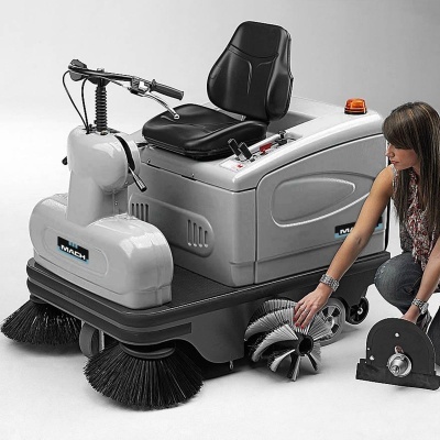 Varredora industrial cinza com assento preto e escovas frontais, mulher segurando peças