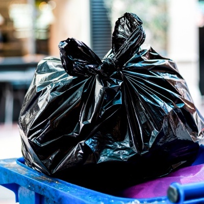 Sacos de lixo pretos amarrados numa lixeira azul com tampa roxa em ambiente urbano