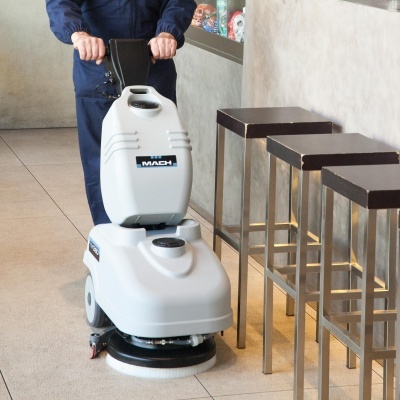 Máquina de limpeza de piso branca MACH em uso num interior com bancos altos metálicos.