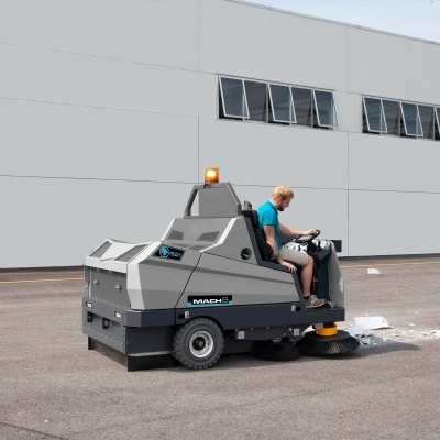 Varredeira industrial cinza com escova amarela operada por uma pessoa em área exterior asfaltada