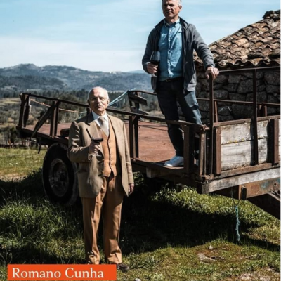 Dois homens com copos de vinho em cenário rural junto a carroça de madeira