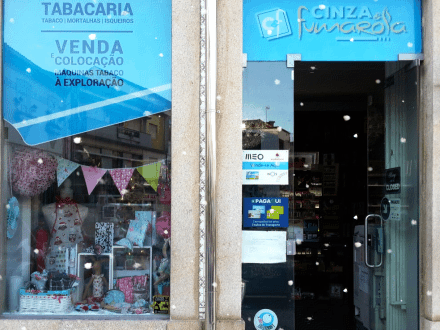 Já é Natal na Cinza Fumarola 🎄 Entrada de loja azul clara com porta de vidro e montra com vários acessórios coloridos e textos de tabacaria