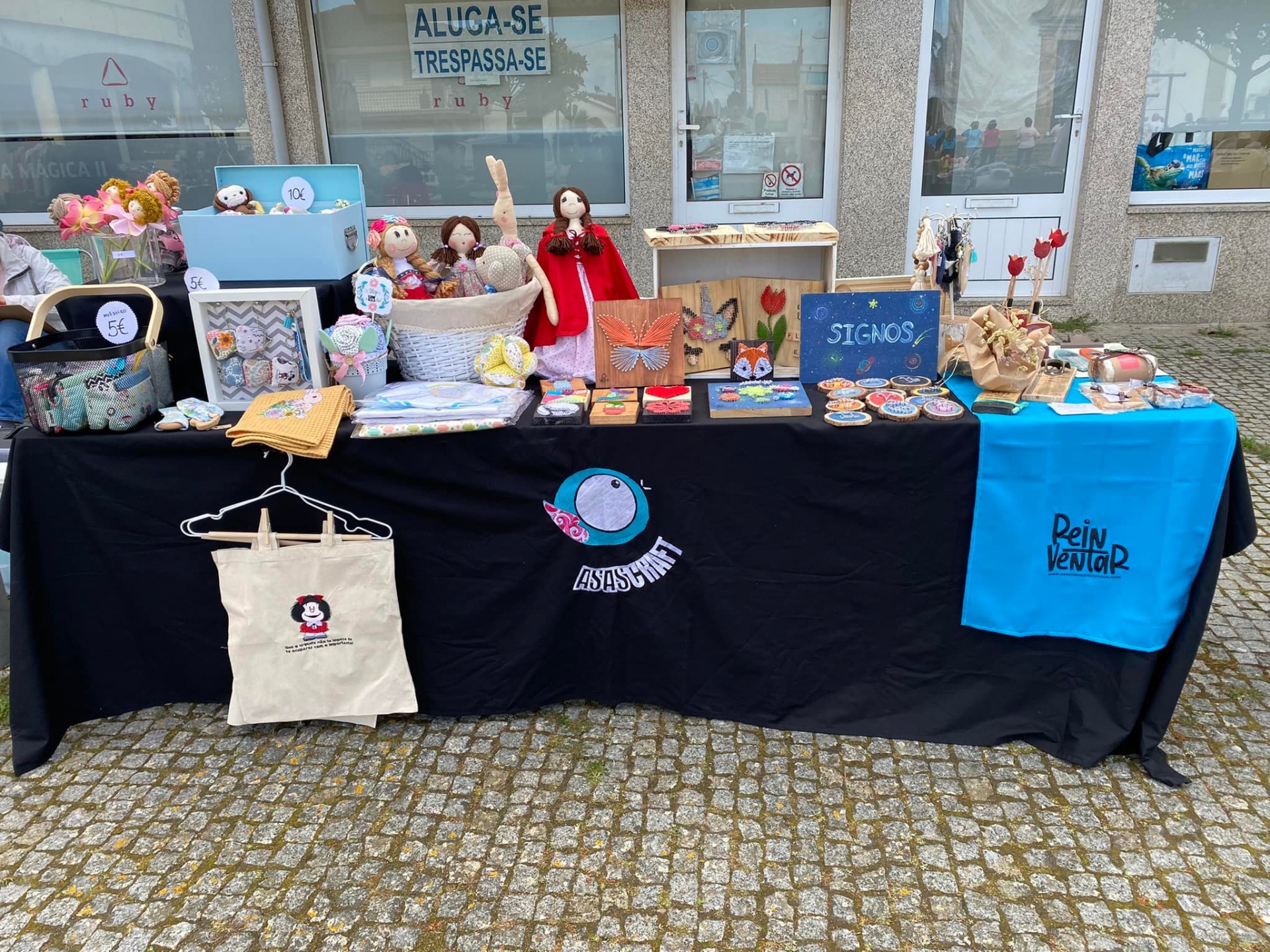 Mesa de venda ao ar livre com produtos artesanais variados incluindo peluches, sacos, quadros e objetos decorativos.
