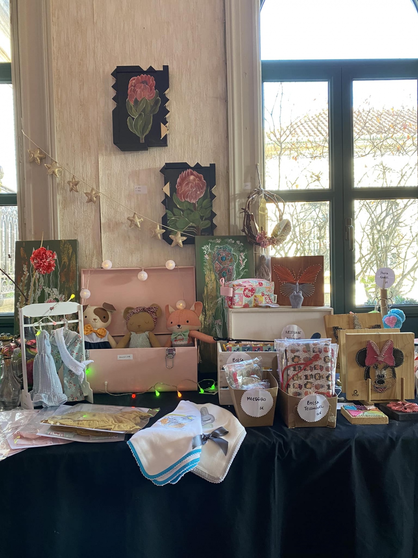 Mesa de artesanato com quadros florais, peluches e sacos com etiquetas junto a janela grande