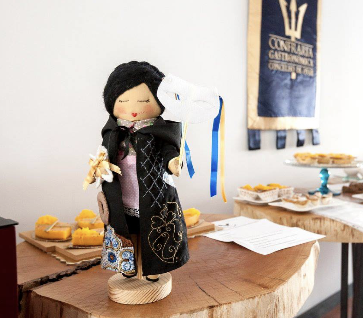 Boneca de pano trajada com traje académico preto sobre mesa de madeira com pastelaria ao fundo