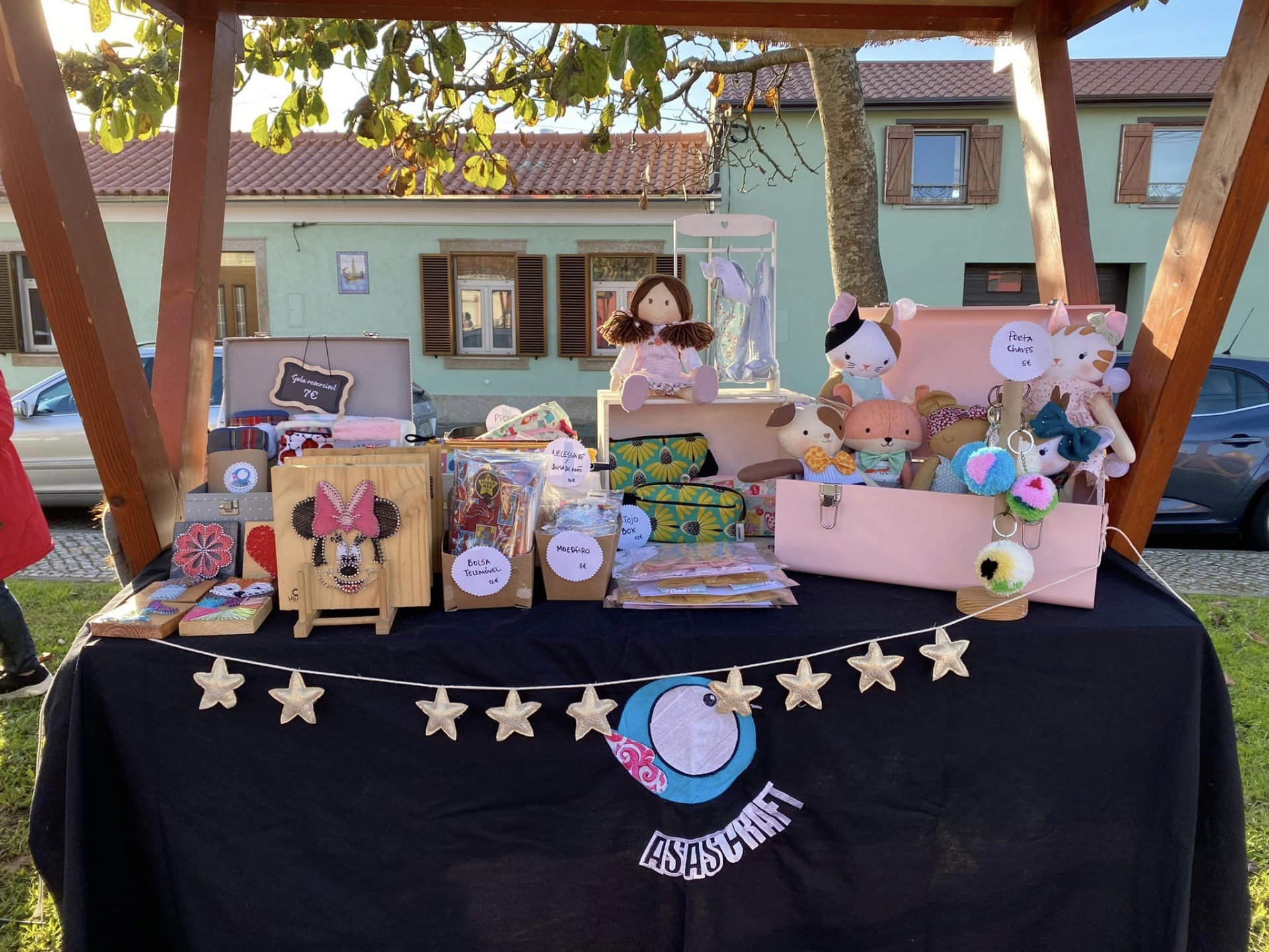 Mesa de venda ao ar livre com peluches coloridos, caixas decoradas e tecidos estampados em ambiente de feirinha