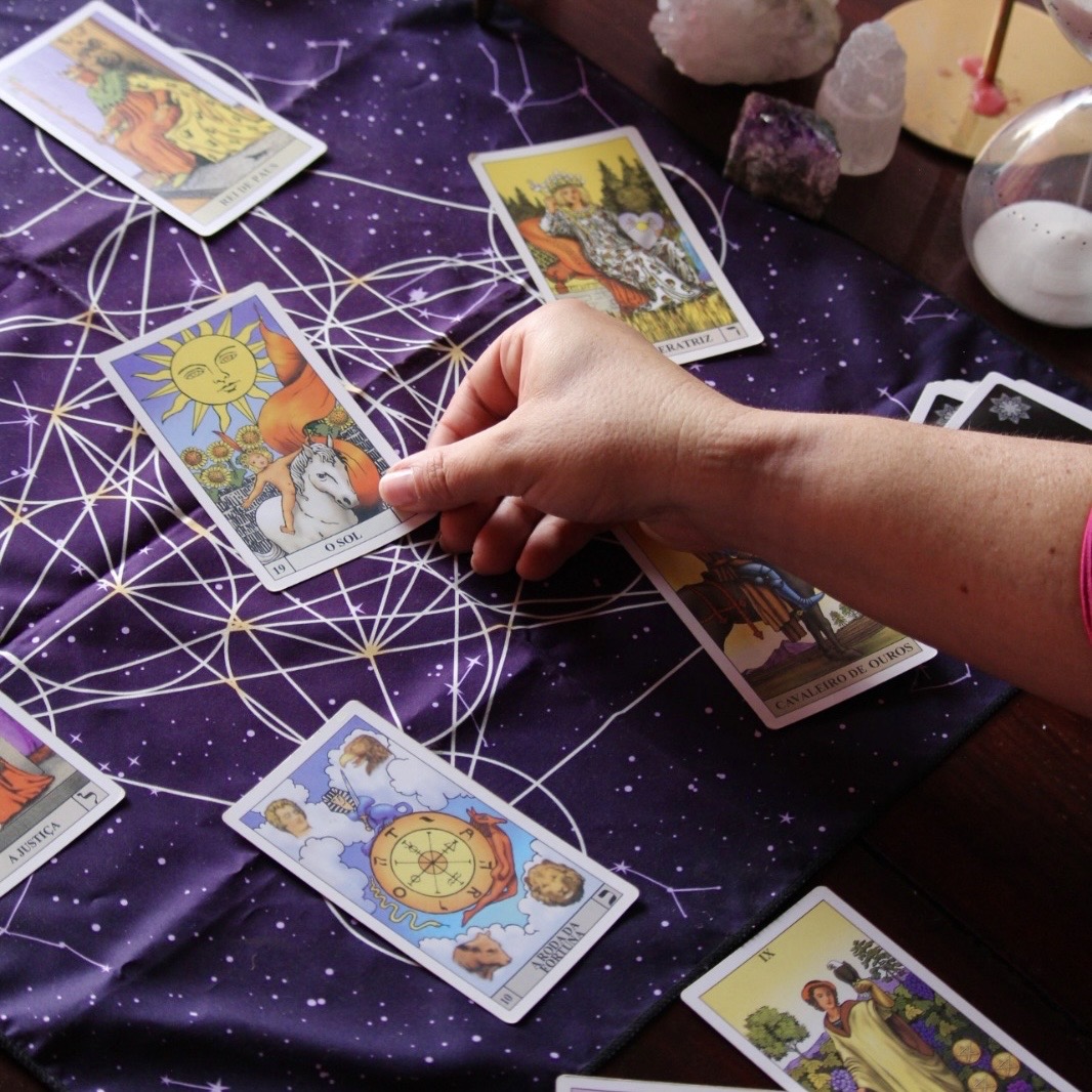 Consulta de tarot terapêutico Tapete com cartas de tarô, mão segurando carta O Sol, ampulheta e cristais