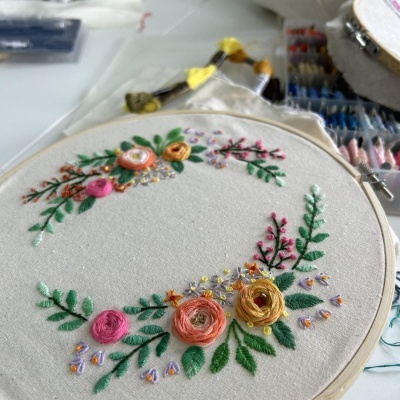 Bastidor de madeira com tecido branco bordado com flores coloridas, agulhas e linhas ao fundo