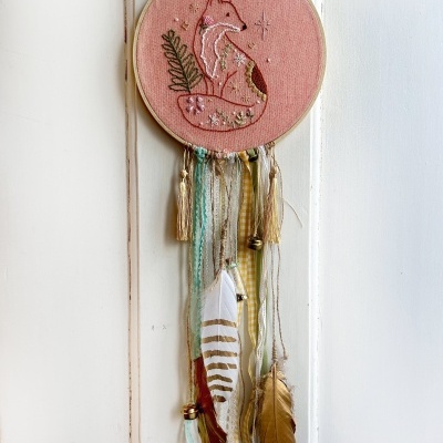 Artesanato decorativo com bordado de raposa em bastidor, fitas e penas penduradas.