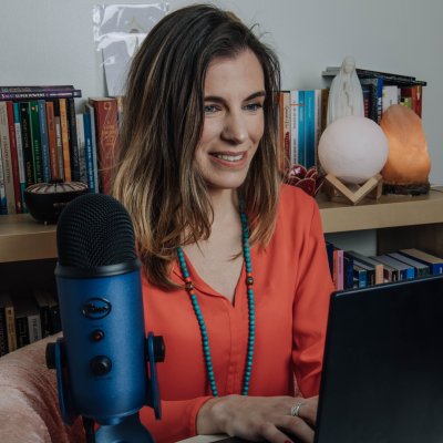 Microfone azul à frente de mulher com camisa vermelha usando computador, com livros e decoração no fundo