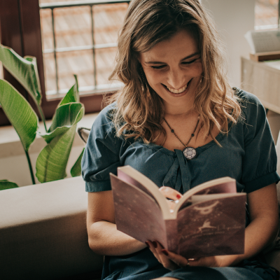 Mulher com vestido azul lendo livro de capa astronómica sentada num sofá com planta ao fundo