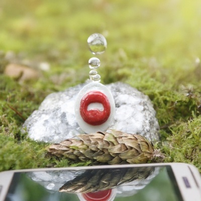 Pingente branco e vermelho sobre pedra com gotas de água e cone de pinheiro sobre musgo, refletido em telemóvel