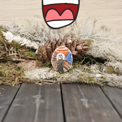 Pedra pintada à mão com desenho colorido sobre pinheiro e musgo em superfície de madeira.