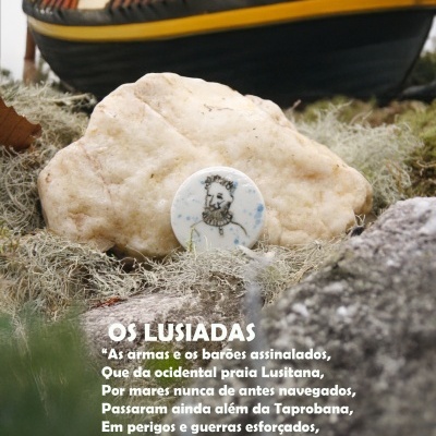 Peça redonda com desenho de busto na pedra com musgo e navio antigo ao fundo com texto OS LUSIADAS