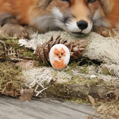 Anel com raposa esculpida branco e laranja sobre musgo com raposa real ao fundo