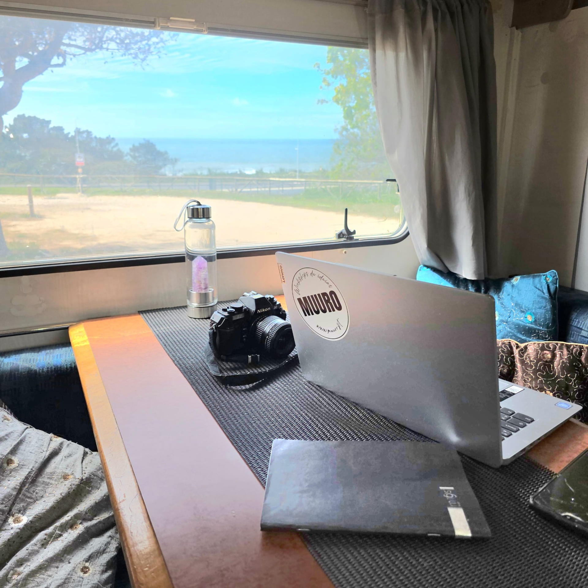 Mesa com portátil MIURO, câmara Nikon, garrafa de vidro, caderno preto numa carrinha com vista para o mar pela janela