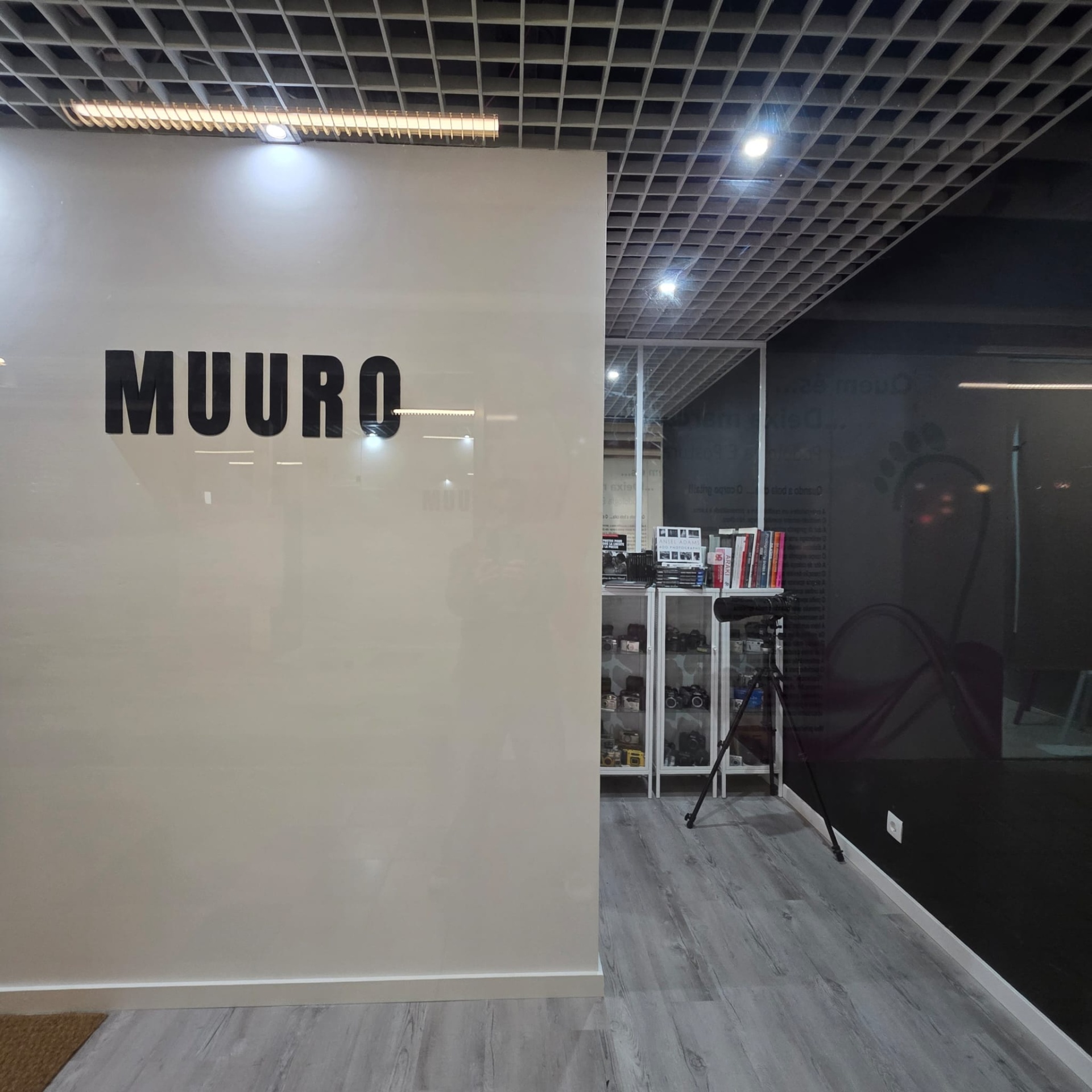 Parede branca com texto MUURO e interior de uma sala com estante e câmaras.