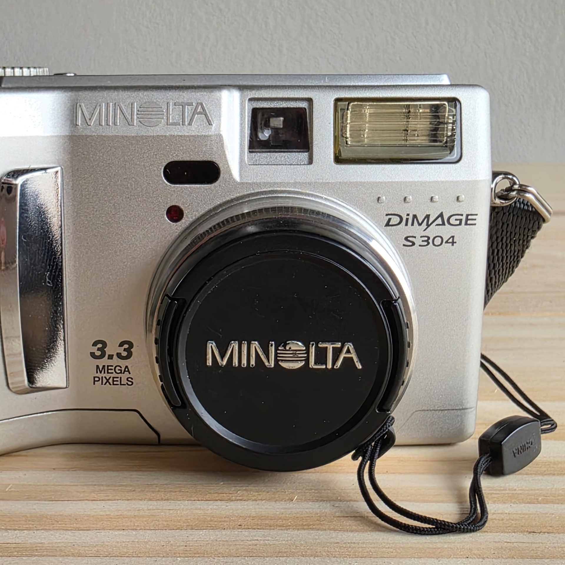 Minolta S304 3,3. Câmara digital MINOLTA prateada com lente, visor e flash sobre tabela de madeira