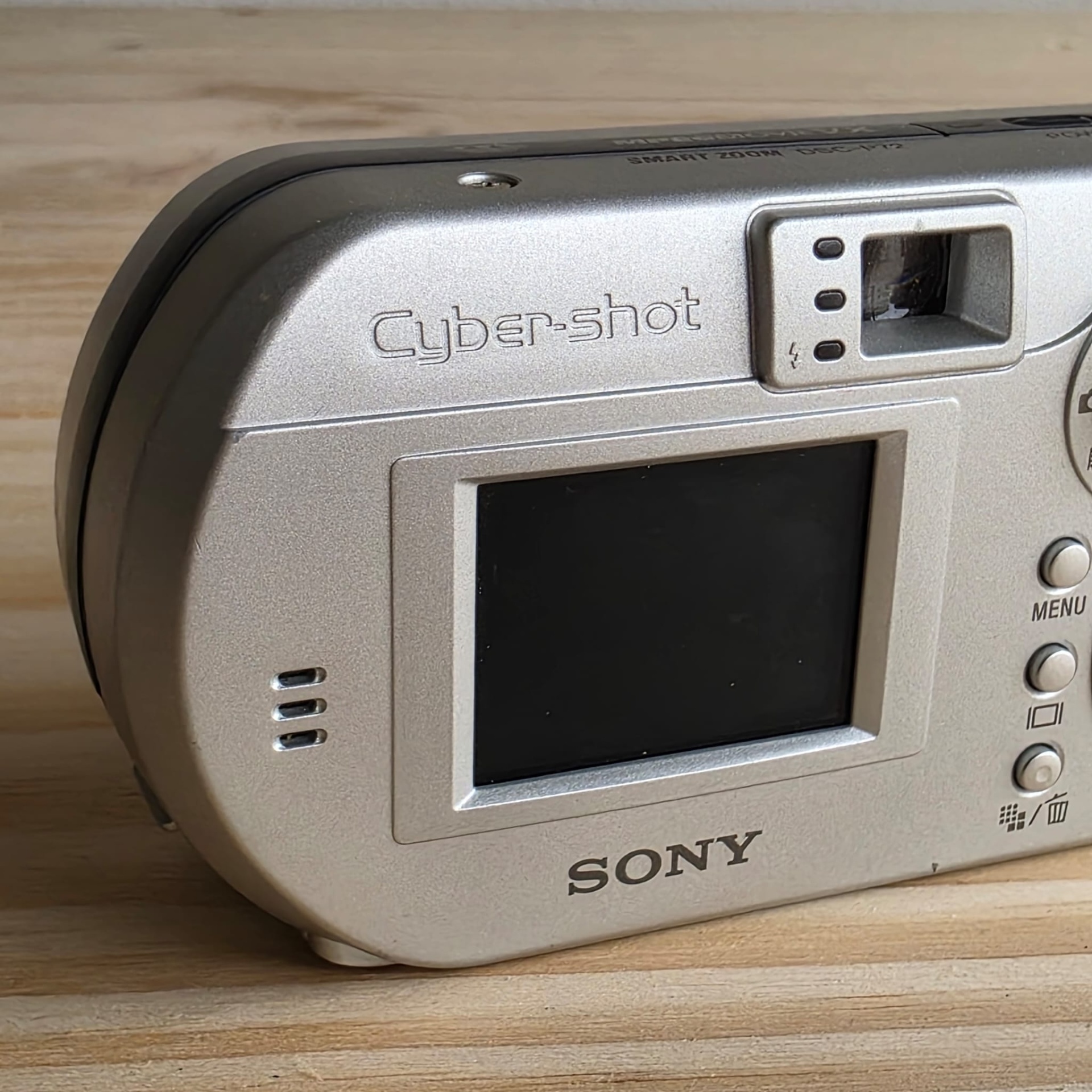 Sony Cyber Shot 3,2 Câmara digital Sony Cyber-shot prateada com ecrã LCD e botões de controlo num fundo de madeira clara