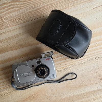 Câmara digital Olympus C-220 Zoom prata com estojo preto sobre superfície de madeira clara.