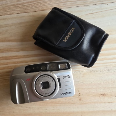 Câmara fotográfica Minolta Zoom 70 prata com estojo preto sobre superfície de madeira clara