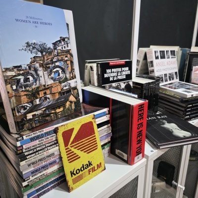Livros de fotografia e artes empilhados, com destaque para capa de livro 28 Millimètres WOMEN ARE HEROES e uma placa amarela Kodak FILM